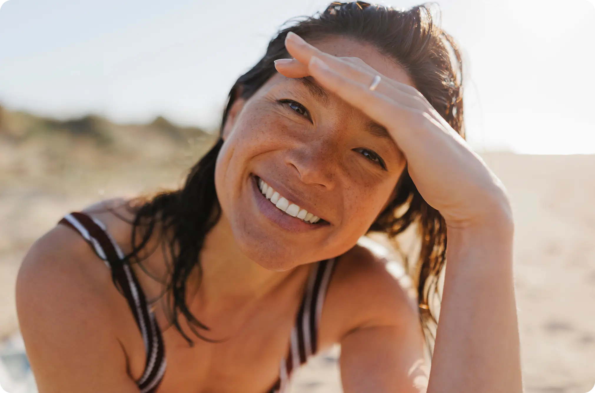 A smiling woman on the beach covering her eyes from the sun
