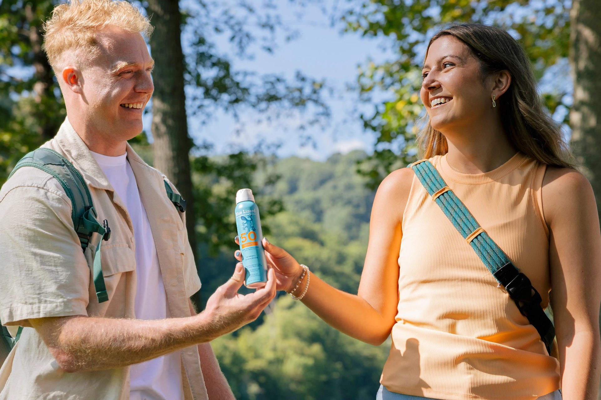 Two people outdoors, one handing Blue Lizard Mineral Sunscreen Spray to the other with trees and mountains in the background.